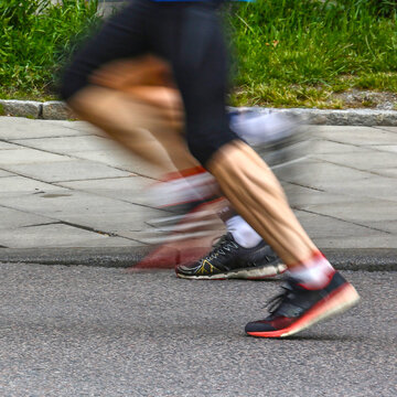 Blurred image of runner legs with motion blur beside urban curb during road race. Square sports image highlights stride, speed and running shoes. Endurance concept