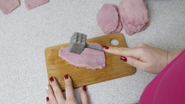 Woman's hands use a metal meat mallet to tenderize a raw pork cutlet on a wooden cutting board. A high-angle top-down shot captures the home cooking process on a kitchen countertop.