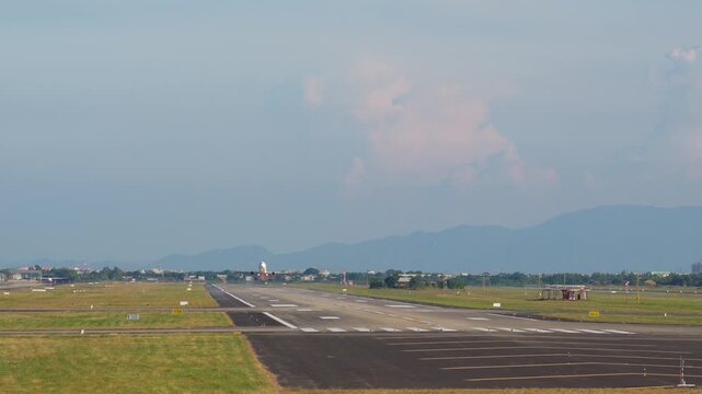 A red and white commercial jet airplane departs from the airport runway, climbing into the sky above grassy fields and distant mountains on a clear day
