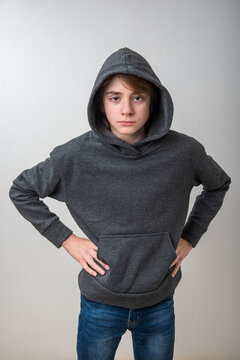 Expressive young boy in a gray hoodie stands with hands on hips against a pale background, projecting a defiant, adolescent attitude and serious, challenging gaze