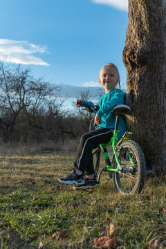 Young child with a happy expression sitting on a bicycle leaning against a tree trunk, enjoying a moment of outdoor play in a natural setting on a sunny day