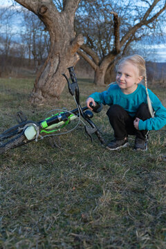 Young blonde girl happily resting beside her bicycle on green grass, enjoying an outdoor activity near trees in the countryside during a quiet afternoon