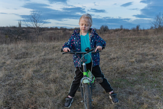 Blonde girl standing on a green bicycle, gripping handlebars and concentrating as she learns to ride in a dry grassy field under an overcast sky, full length portrait