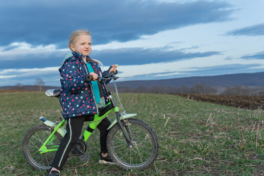 Blonde child wearing a colorful jacket and sitting on a green bike in a rural field under a cloudy sky, looking ahead with a sense of wonder and freedom