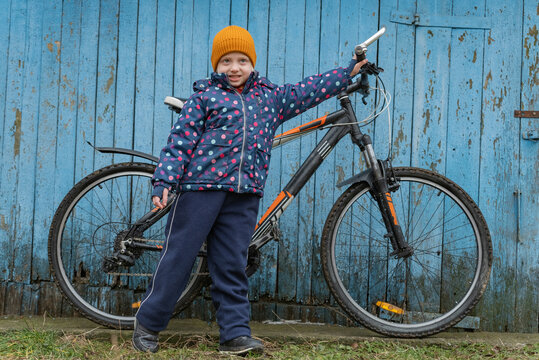 Happy girl in a warm hat and jacket standing with her bicycle beside a weathered blue wooden wall, smiling and enjoying a cold outdoor bike break during winter recreation