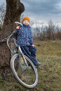 Young girl in a yellow hat and polka dot jacket sits by a tree holding her bicycle handlebar, smiling happily while enjoying outdoor play and carefree childhood in a park like setting