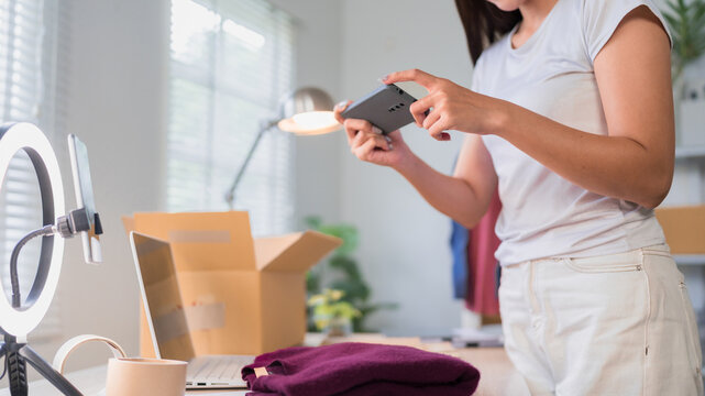 Young Asian woman entrepreneur using smartphone to manage online business while preparing product parcels in home office