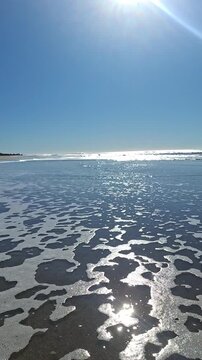 Vertical backlit medium shot of glistening ocean water and sea foam on a sandy beach, sparkling sun reflection on wet shore, clean coastal loopable background.