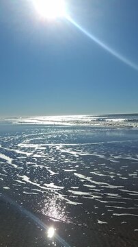 Vertical medium shot of a beautiful sandy beach with ocean waves and sea foam under a bright sun lens flare, sparkling sunlight reflecting on water surface, coastal nature scenery.