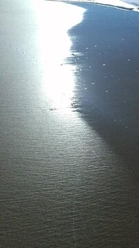 Vertical panning shot revealing a beautiful sandy beach coastline with sparkling sunlight on the sea surface, ocean waves washing onto wet shore, summer nature background.