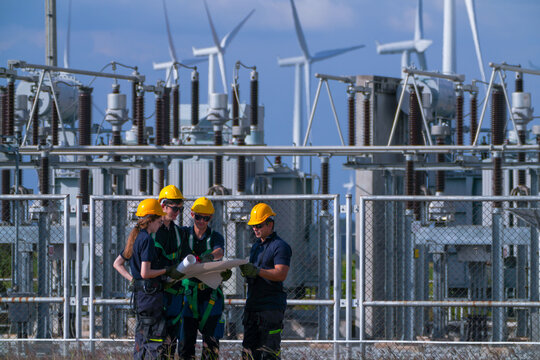 A commissioning team in navy suits and yellow hard hats point along the busbars while comparing the drawing inside the wind farm grid substation in crisp blue sky daylight at a wind power.