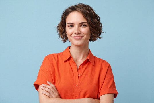 Portrait of a smiling young woman with short wavy hair wearing an orange shirt standing against a light blue background