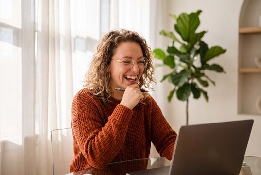 Happy young woman with curly hair laughing while working on a laptop at home
