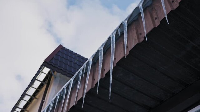 Long, sharp icicles hang in a row from the eaves of a house roof. White clouds move quickly across the sky in a time-lapse shot, indicating the passage of time.