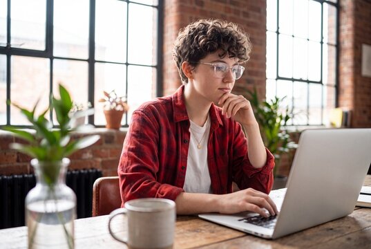 Young person with curly hair and glasses working on a laptop in a sunlit industrial office space