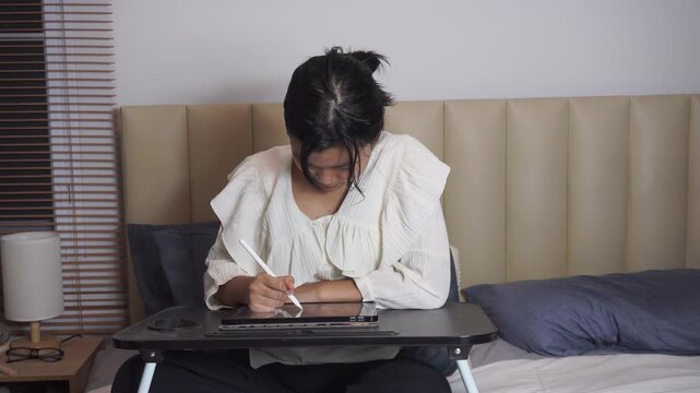 Teenage girl using tablet on bed at night. happy Asian student is drawing with stylus pen inside bedroom during her Gen Z lifestyle creative art hobby remote freelance job