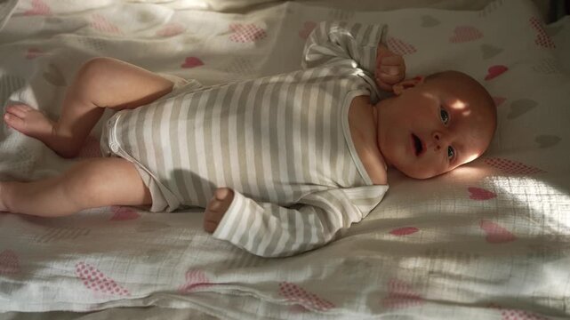 Adorable newborn baby boy in a striped bodysuit lying on a bed with a heart patterned sheet. The infant is looking around, yawning, and then starts to cry in the morning sunlight