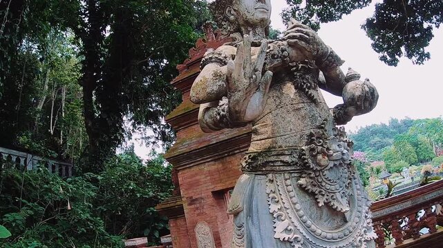 Foreground stone guardian statue stands beside red temple arch in lush tropical garden, with smaller matching figure, carved stairway, dense foliage, and tall trees creating serene exotic setting in