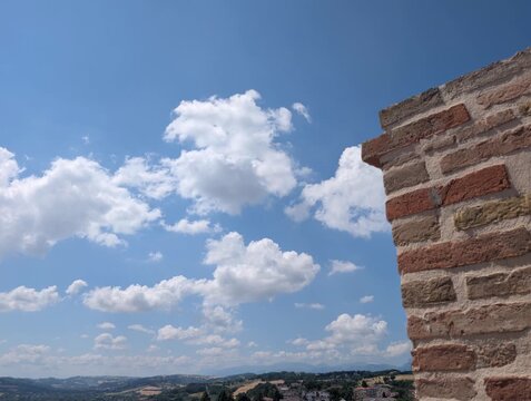 Un muro in mattoni emerge in primo piano sotto un cielo limpido, mentre sullo sfondo si apre un paesaggio di colline e piccoli edifici