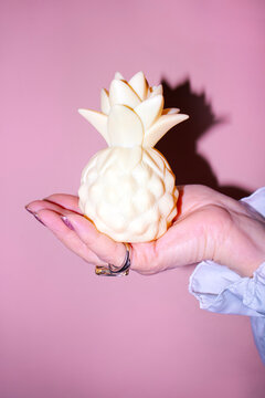Hand holding a decorative pineapple-shaped candle in soft yellow color against a pastel pink background, showcasing intricate details of the design