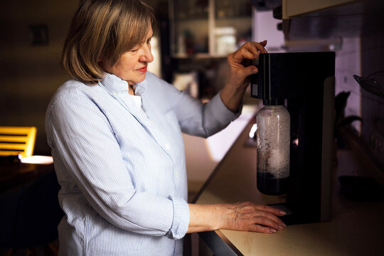 Senior woman in blue striped shirt using a soda maker in a modern kitchen, with a countertop and kitchen appliances visible in the background