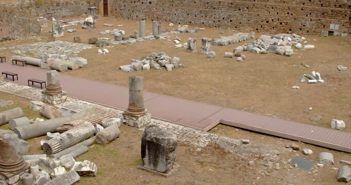 An archaeological courtyard displays broken marble columns, stone artifacts, and historic brick walls, featuring a modern pedestrian walkway designed to allow tourists to view the preserved remnants.