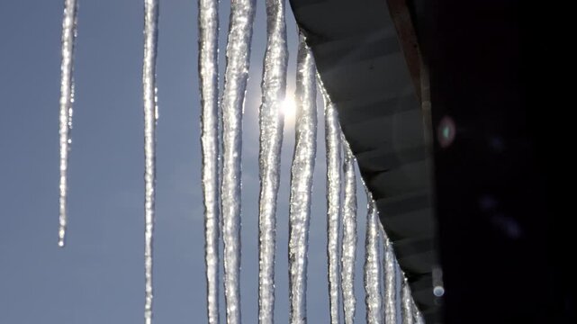 Transparent icicles hang from a metal roof edge, dripping water as the bright sun shines through them against a blue sky. The scene represents the spring thaw and seasonal change.