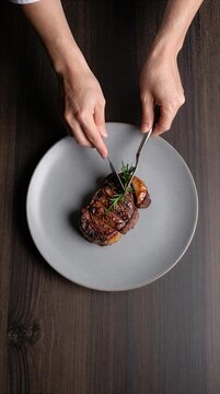 Person cuts a piece of steak on a round gray plate with hands holding knife and fork