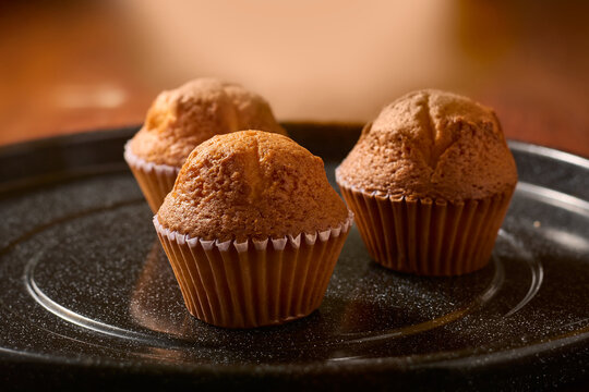 three freshly baked muffins on black textured plate, golden brown sponge cakes in paper cups, side view with soft focus and copy space, warm homemade dessert