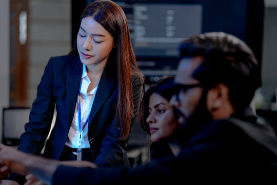 Two diverse professionals in suits collaborate in a modern tech office. One uses a stylus on a tablet while the other observes, analyzing complex data on multiple screens.