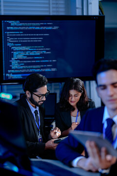 An AI research team brainstorms late night. Innovators discuss machine learning model architecture using a tablet while reviewing training data code displayed on a large screen.