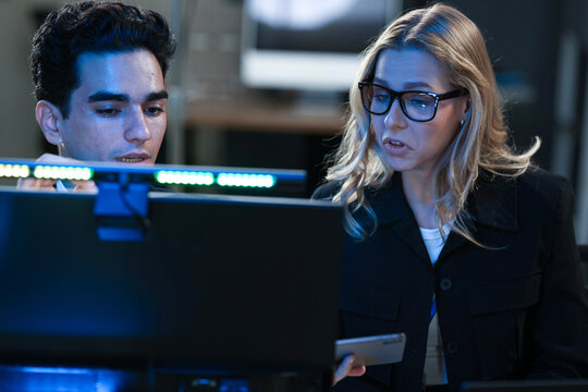 Dynamic tech team collaborates late night. Young Asian woman points at screen explaining project details to male colleague reviewing documents in modern hub workspace.