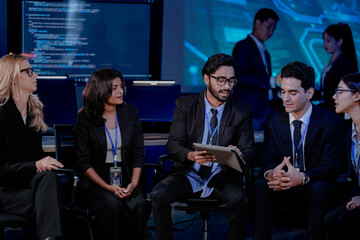 Obraz premium A large, diverse team of business professionals in a group meeting. An Indian man with a tablet leads the discussion with Asian and Caucasian colleagues in a high-tech office.