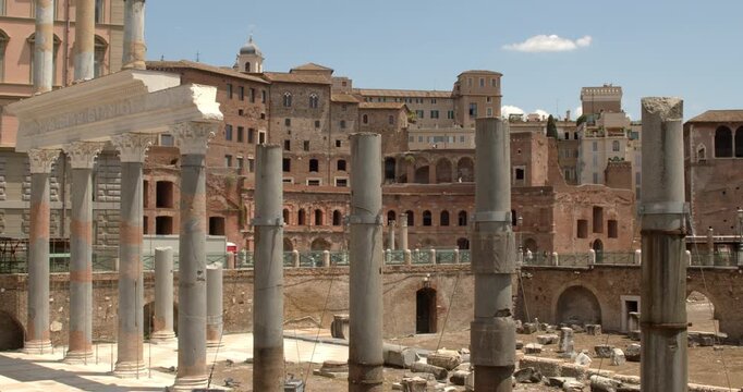 A closeup view of the Trajan's Forum archaeological site in Rome, Italy, displaying weathered granite columns standing among ancient stone fragments, brick structures, and historical buildings.