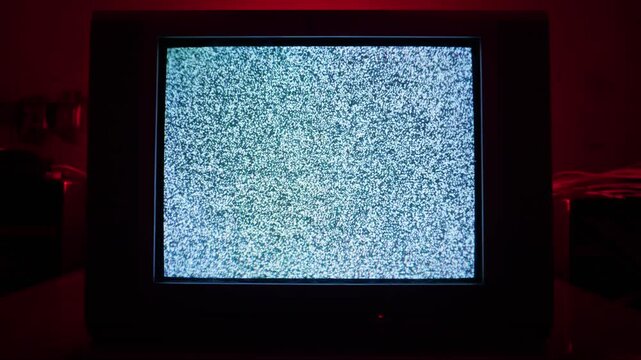 Vintage CRT television displays flickering static noise in a dark room with red ambient light. The eerie interruption portrays a concept of technological glitch or suspenseful horror.