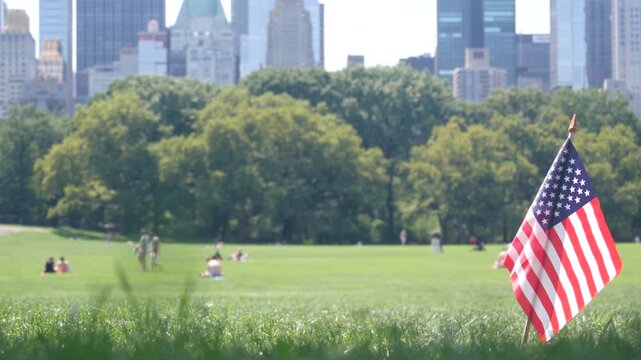 American flag on green grass in Central Park, New York, USA. Sheep Meadow, high-rise buildings and people. Skyline cityscape, Manhattan Midtown skyscrapers. United States symbol and business district.