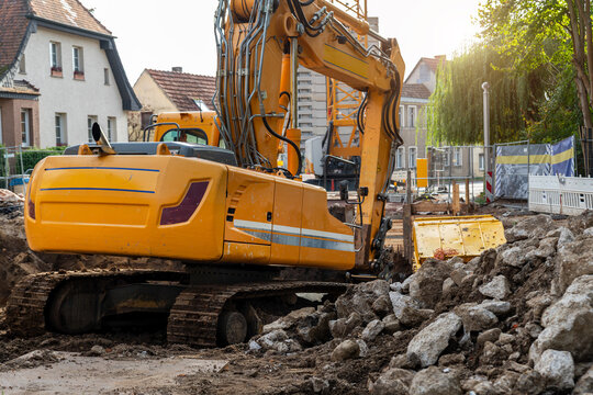 Yellow excavator works on urban construction site beside rubble, soil, concrete, fences, and residential houses. Hydraulic boom, crawler tracks, and debris show roadwork, renovation, infrastructure