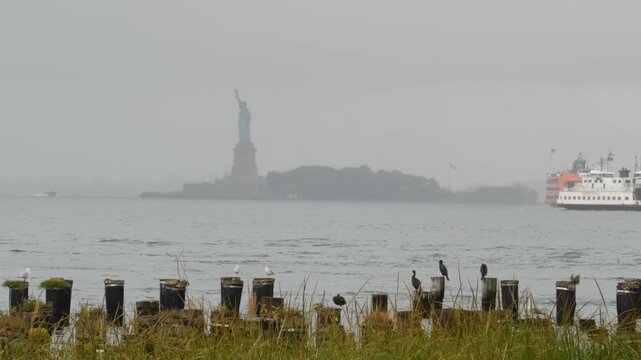 Statue of Liberty on Liberty Island from Brooklyn, New York City, United States. Ferry to Staten Island and birds on old wooden pier piles remains. Rainy foggy day weather. Orange boat water transport
