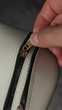 A hand grasps a silver metal zipper pull and slides it along the black teeth of a white leather bag in a close-up shot. Concept of security, organization, and fashion accessories.