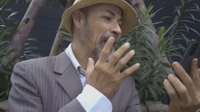 Inspiring mature man Wear a hat, wear a suit nice beard talking Serious steady, with a smile, in encouraging the other person to smartphone camera, making OK sign and thumbs up gesture in garden.