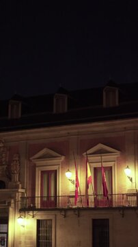 Vertical night view of a historic Spanish government building with illuminated stone facade, balconies, flags, and classical architectural details under a dark night sky.