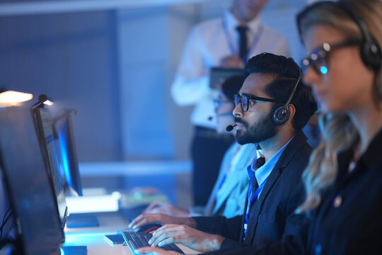 Operators wearing headsets work at computers in a dark modern call center monitoring data on glowing screens at night.