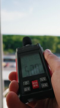 A person holds a digital sound level meter measuring a noise level of 49.8 decibels near an open window overlooking a green area. Concept of environmental noise monitoring and urban acoustics