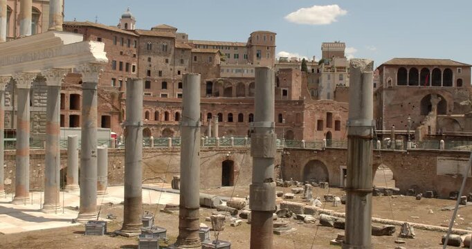 A closeup view of the Trajan's Forum archaeological site in Rome, Italy, displaying weathered granite columns standing among ancient stone fragments, brick structures, and historical arches.