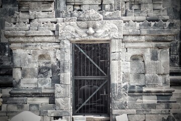 Fototapeta premium Ancient stone entrance of Candi Ngawen temple featuring Kalamakara carving and gated chamber with yellow leaves foreground, Muntilan, Central Java, Indonesia.