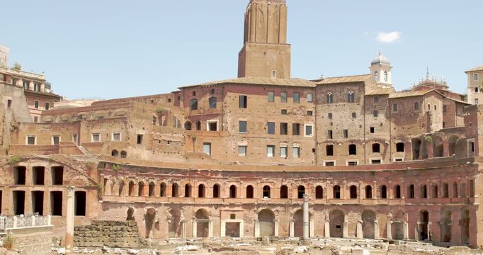 Panorama of Trajan's Market in Rome, Italy, showcasing the semi-circular ancient brick structure and multi-level ruins. A medieval stone tower rises in the background under a pale summer sky.