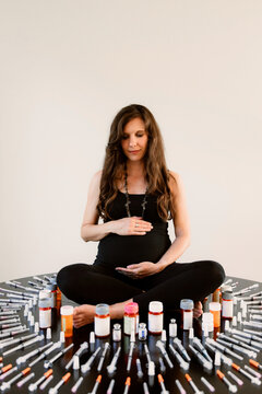 Pregnant woman sitting cross-legged surrounded by IVF syringes and medicine bottles
