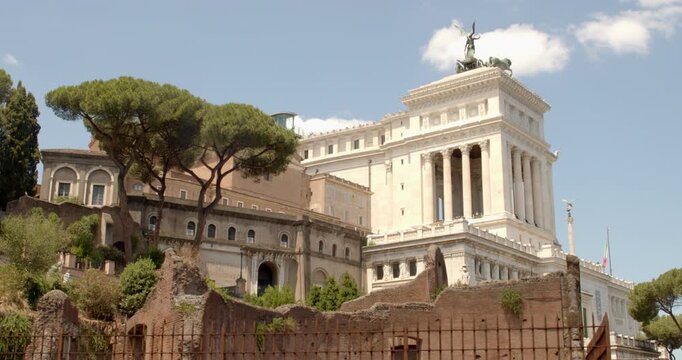 A daytime shot in Rome, Italy, showcasing the striking contrast between the ancient stone ruins on the Capitoline hill and the massive, white marble Vittoriano monument rising up in the background.