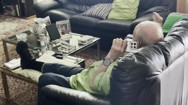 Abuelo usando smartphone viejo con tel&eacute;fono hablando en sal&oacute;n anciano sentado hablando por el m&oacute;vil y gesticulando con la mano tercera edad y celular en sof&aacute; v&iacute;deo dom&eacute;stico gestual expresivo