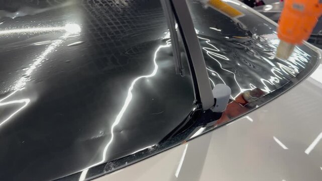 Professional car window tinting process: technician prepares the glass, applies tint film, uses a heat gun to shape it on curved surfaces, and smooths out air bubbles with a squeegee. 